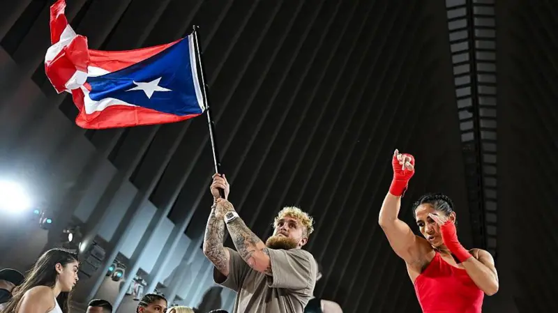 Jake Paul waves a Puerto Rico flag and stands next to Amanda Serrano who is throwing a punch