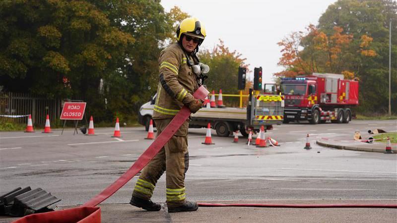 A male firefighter reeling up a red hose from the floor with his hands. He wears fire-resistant clothing, a yellow helmet and black sunglasses. He is stood up with his body moving away from the camera but his head is looking back in the direction of the camera and the red hose he is reeling up. Behind him are traffic cones, a road closed sign, temporary traffic lights and a fire engine. 