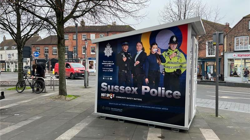 A police box in Chichester City Centre on a pavement with an advert for Sussex Police showing various uniformed and non uniformed officers