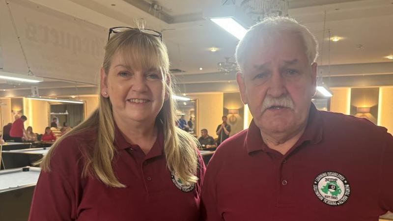 A blond haired woman and a white haired man stand beside one another. She is smiling and has glasses on her head. He has a white moustache. Both are wearing red polo shirts.