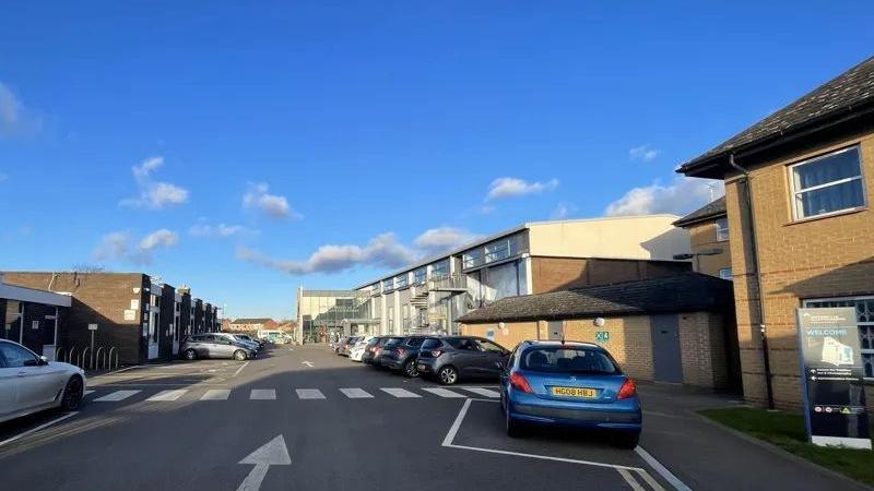 A view of the current campus, with a few houses and cars parked on a road going through them. The sky is blue.