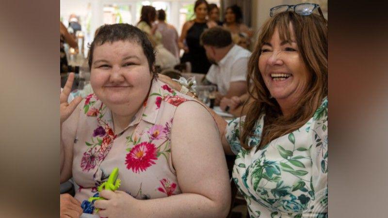 Mum Rebecca on the left. She has brown hair and is wearing a beige dress with green leaves on. Her daughter Alice is on the left. She is wearing a pink blouse shirt with purple and pink flowers on. She has shorter hair. Both are at a wedding.