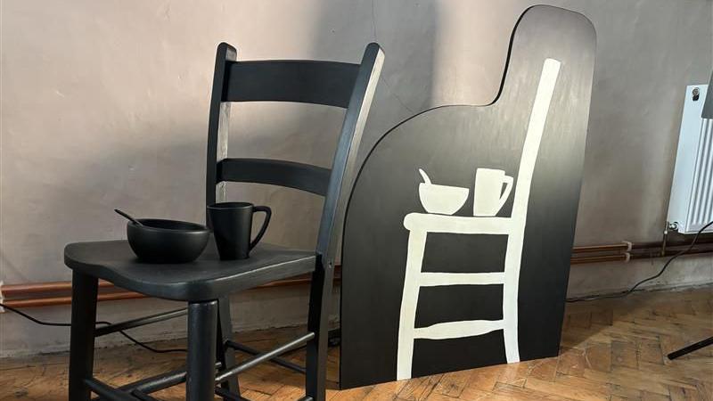 A black chair with a black bowl and mug stand in fron tof a silhouette of a white chair against a black background in the artist's exhibition 