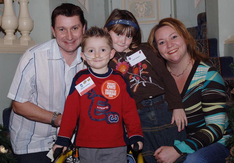The family are posing besides some stairs. Chris has a white shirt with blue stripes, Matthew has a red top and has a feeding tube and a badge reading Children of Courage, Laura is wearing a brown top and blue denim skirt and Lisa has a pale blue, yellow and brown striped top.