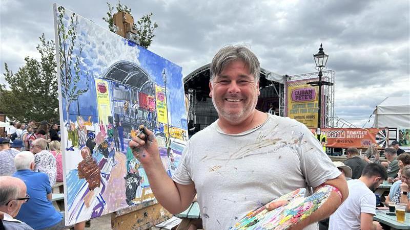 A man with short grey hair, wearing a grey t-shirt and holding a board with paint on in his left hand. He is holding a paintbrush in his right hand and is holding it up to a painting. He is smiling at the camera and is stood in a crowd of people. 
