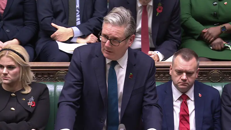 Keir Starmer at the despatch box in the House of Commons. He is wearing a blue suit and has a poppy on his lapel. He is flanked by two other MP who are sitting on the green benches behind him.