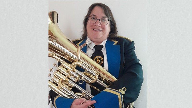 Rose in full black brass band suit with gold and blue trim. She is carrying a tuba.
