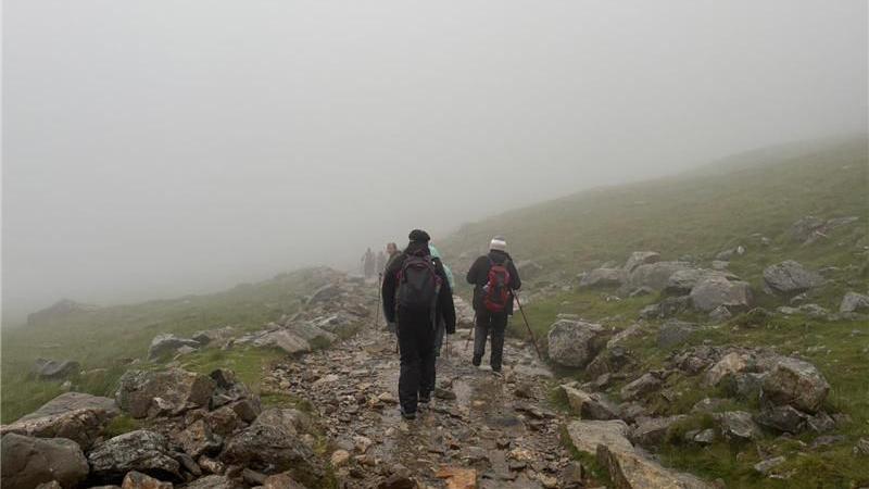 Several people are hiking on a rocky mountain path. There is dense fog all around them. They are wearing hiking gear and have hiking poles. You can make out green grass and grey rocks either side of the path