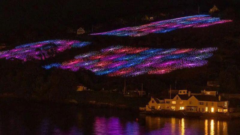 A field filled with lights. There are squares of purple, pink, blue and red filling the field. To the bottom right there is a house.