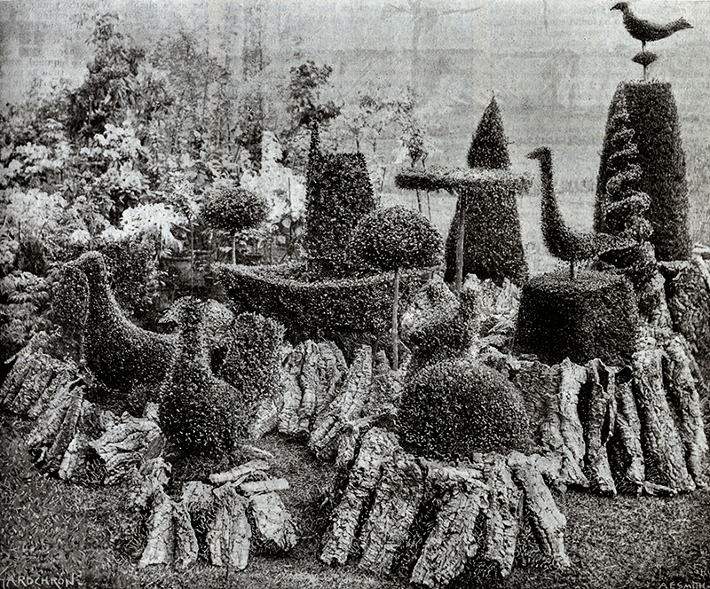 a photo of some topiary in different shapes set out in a circle. The topiary bushes are shaped into birds, circular mounds, vertical spirals and there is a wheelbarrow one in the middle. They are positioned on top of what looks like sawn off tree trunks.