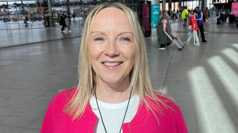 Ms Doyle is smiling at the camera. She has long blonde hair and is wearing a bright pink zip up jacket and white t-shirt.
