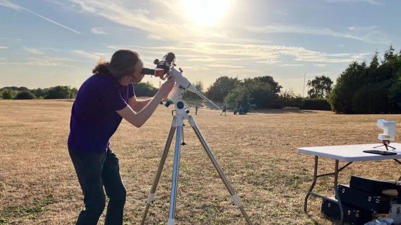 A man in a purple T-shirt and black trousers peers through a telescope.