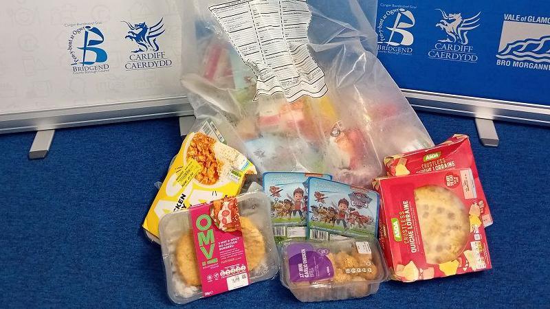 A bag of packaged food with some lying in front of it. A chicken curry and rice ready meal, a chicken burger with a red label, two red boxes of quiches, garlic chicken bites and paw patrol snack bites.