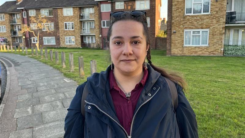 Eszther Kadar, a middle-aged woman, poses for a photograph in Rosewood Road. She wears a black jacket over a red shirt, with sunglasses pushed up above her head.