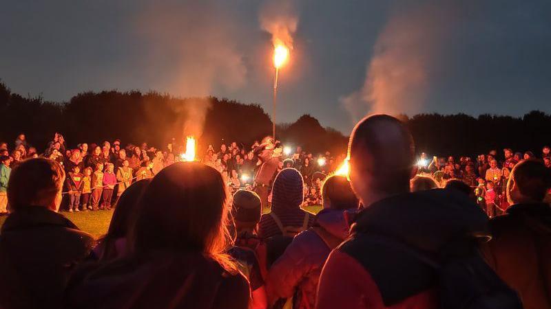 In darkness, a large crown of people gather, lanterns light up their faces