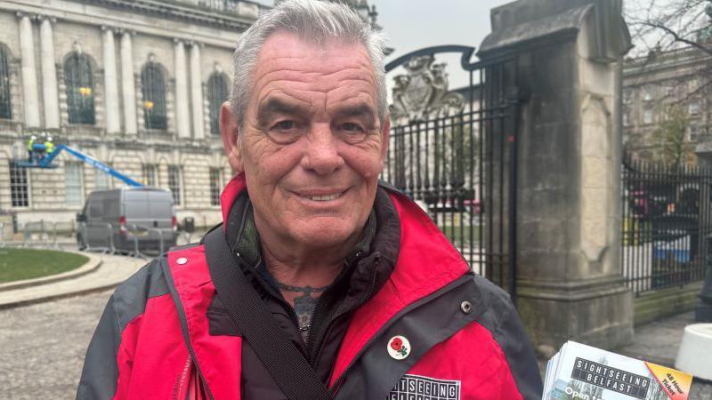 A manager of Belfast sightseeing standing in front of City Hall Belfast. He has grey hair and tattoos on his chest. He is wearing a red and black coat. The coat is labelled Sightseeing Belfast. The coat has a poppy badge. He has leaflets in his hand standing in front of the city hall. 