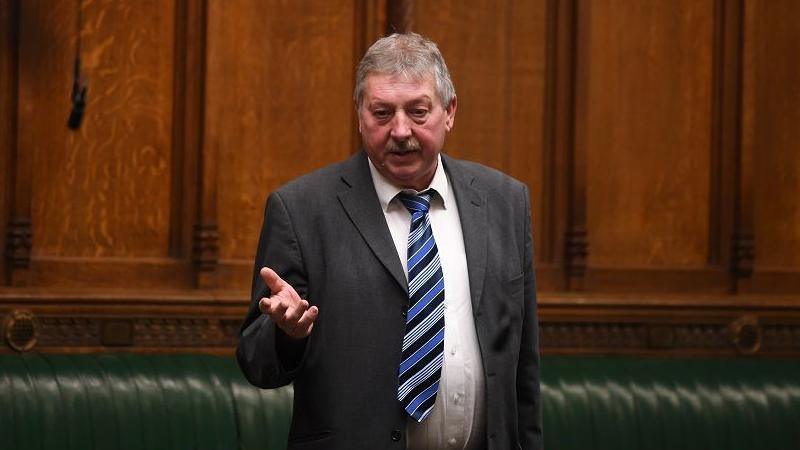 Sammy Wilson in parliament wearing a grey suit, white shirt and blue/black diagonal striped tie. He has short grey/light hair and a grey/white moustache. He is speaking and has his hand elevated in front of him.