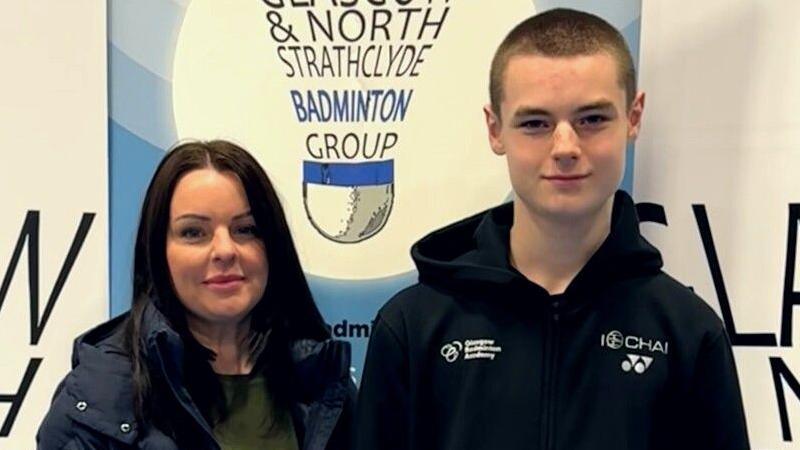 Michelle Junnor has long dark brown hair and smiles at the camera. She is wearing a green top with a black puffa jacket on top. Next to her is her son Charlie. He is taller than her and wears a black hoodie. Behind them a sign reads "Glasgow & North Strathclyde Badminton Group".