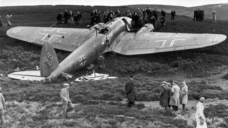 a crashed German WWII aircraft with visible damage and a swastika on the tail. Several people are gathered around the wreckage.