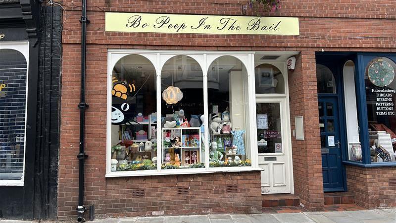 The outside of a shop window with three arched windows and a white door to the right. A black cat with an orange hat logo is peeking out from the left and there are several teddies on shelves in the window on display. At the top of the windows, a yellow and black sign reads "Bo Peep In The Bail".