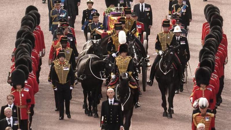 The coffin of Queen Elizabeth II, draped in the Royal Standard with the Imperial State Crown placed on top, being carried along Whitehall on a horse-drawn gun carriage. 