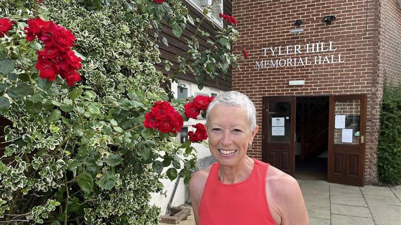 A woman with short hair and a red vest top stands outside Tyler Hill Memorial Hall next to a rose bush