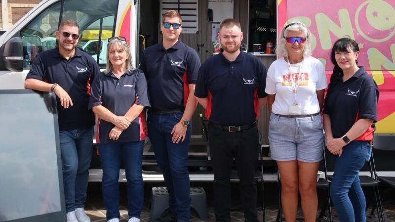 Six people, three men, and three women, standing outside a van, looking at the camera. The van door is open behind them and they are mostly smiling.