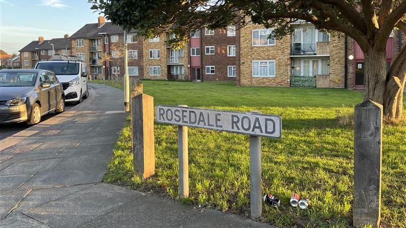 The concrete road sign for Rosedale Road, with several parked cars in the background.