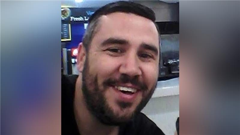 A man with short black hair and a black beard smiles at the camera. He is wearing a black shirt.