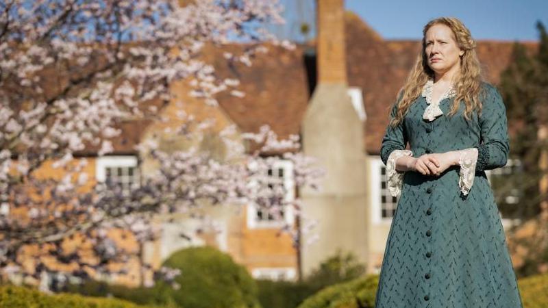 A woman in Edwardian dress and long blonde wavy hair stands in the foreground of a red brick medieval mansion. There is also a blossoming tree in the background.