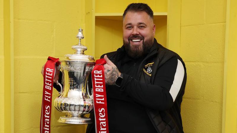 Harborough Town manager Mitch Austin smiling whilst holding the FA Cup aloft.