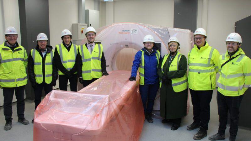 Eight people in high-vis jackets and white hard hats stand around a full-body diagnostic scanner that is wrapped in pink plastic.