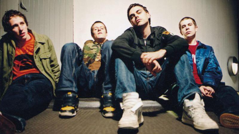 Hard-Fi, shot from a low angle, sit on a plain carpeted floor, wearing early-2000s combat gear-style fashion