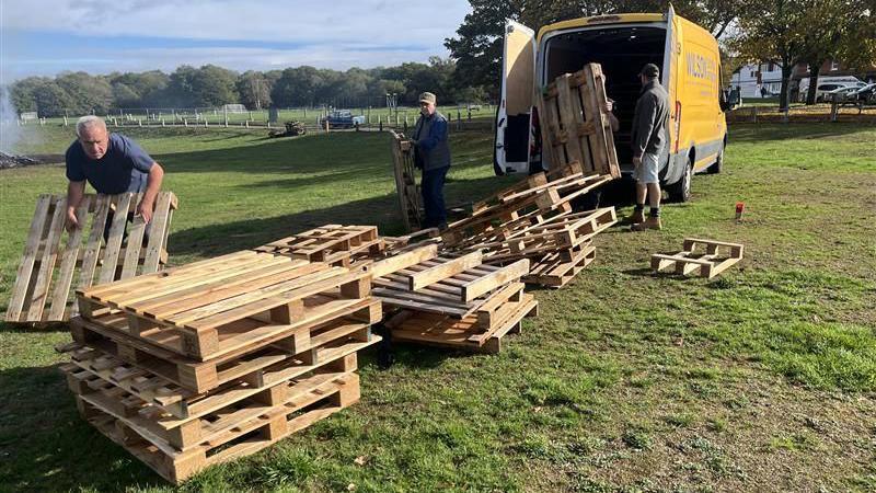 Three men pulling wooden pallets from a yellow van. The pallets are being stacked on grass.