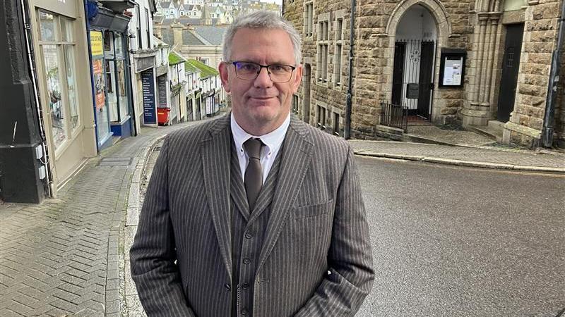 Johnnie Wells smiles as he stands on the pavement of the narrow road which has a sharp bend in it and leads down hill. He is wearing a brown pin striped three-piece suit with a brown tie. He has short grey hair and is wearing a pair of glasses.