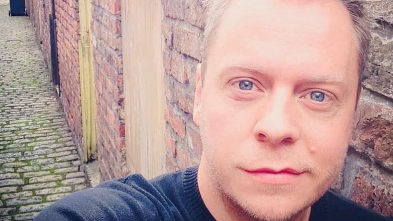 A selfie of a smiling Lee Salisbury in an alleyway. He has blue eyes, blond hair and blond stubble along his jawline, upper and lower lip. His hairline appears to be receding. 