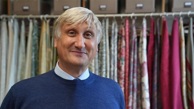A man smiles at the camera. He has grey hair and is wearing a blue jumper with a blue shirt underneath. Behind him, long drapes of silk are hung up on a curtain pole.