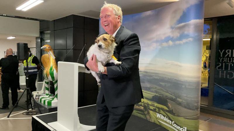 Image of Brian Mathew. He has white hair and is wearing a black suit. He is smiling and holding his Heidi, his brown and white 