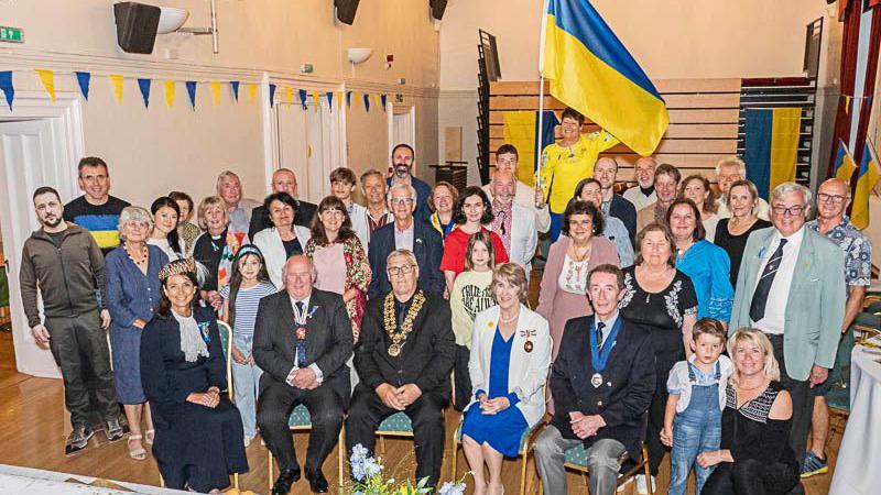 A large group of people sat in hall with Ukrainian flags and bunting