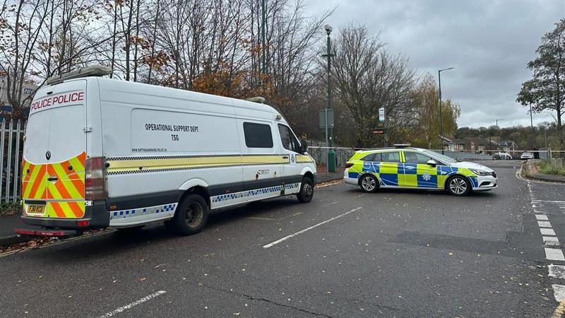 A large white police van parked next to a police car on a two-lane road. A cordon can be seen horizontally at the back of the image.