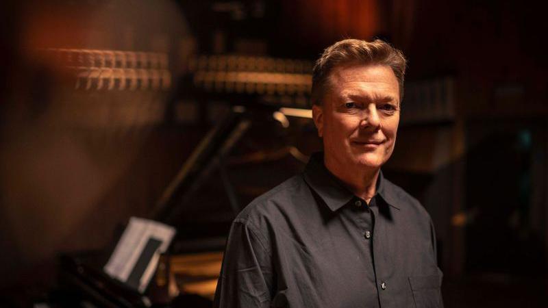 Toby Spence, a man wearing a dark shirt, stands in front of a black grand piano looking into the camera