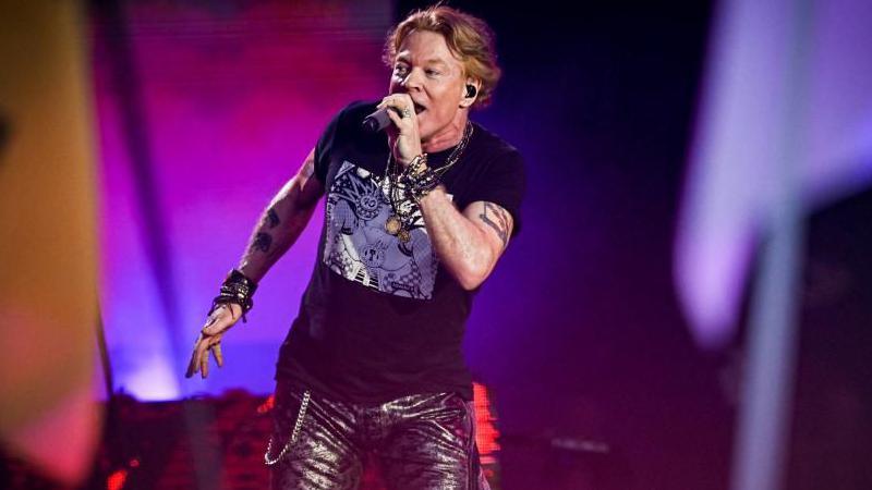 A rock singer on the Pyramid Stage at Glastonbury. He has reddish brown hair and is wearing black leather trousers and bright purple lights illuminate the stage. He has tattoos on both of his arms and is singing into a microphone.