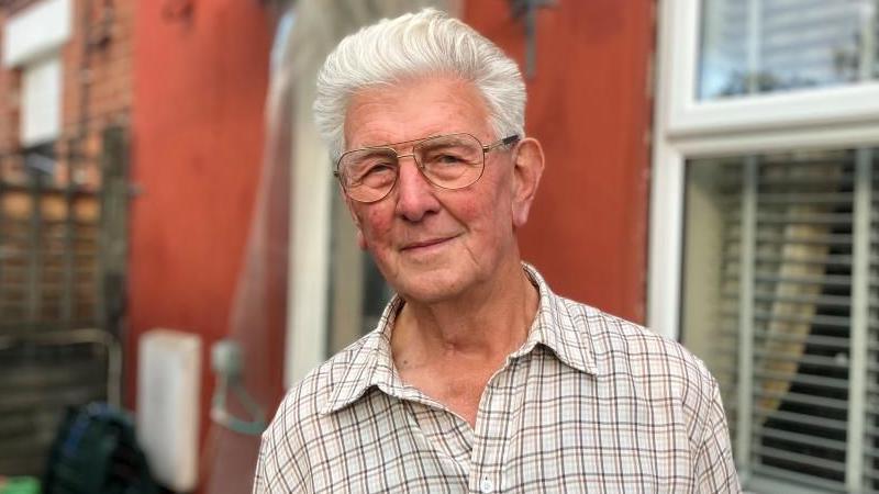Image of David. He is stood in front of his home. He is wearing glasses, a chequered shirt and has white hair.