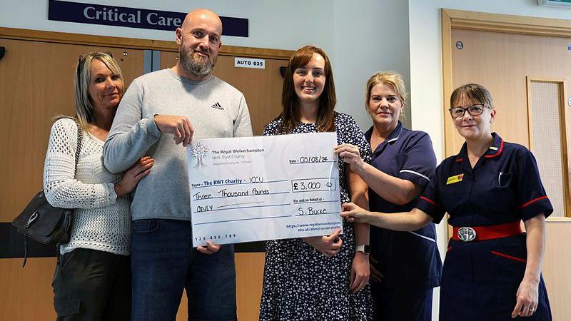 Simon Burke stands second from left with a beard and shaved head, wearing a grey sweatshirt. He is presenting a cheque. His wife is on one side and three fundraising and clinical staff from New Cross are on the other 