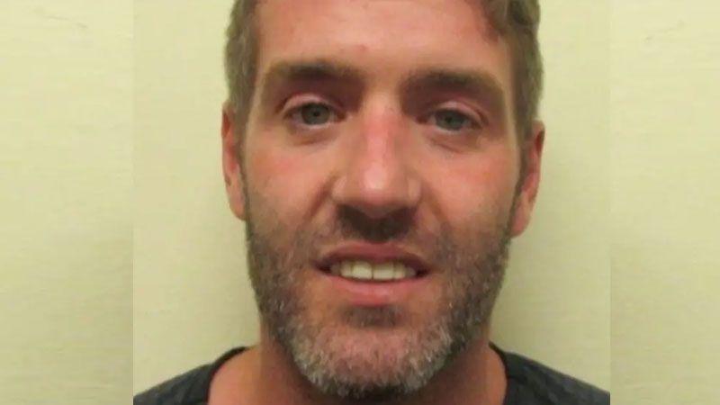 A police mugshot of a smirking Jamie Rothwell, who has dark stubble tinged with grey and light brown.