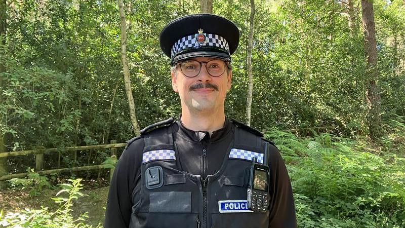 A policeman in the countryside. He has a moustache and is smiling.