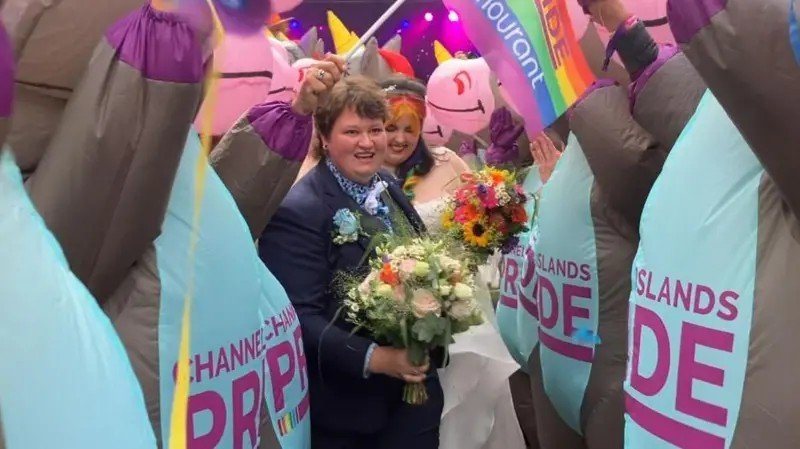 Two smiling women carrying flowers, one in white wedding dress and another in a blue suit. They walk through a procession of people dressed in blue and grey donkey-corns with the words Channel Islands Pride on the bellies in purple writing.