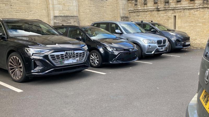 Four cars in a car park. Three are black, one is grey. There's another car to the right of the picture, just out of shot.