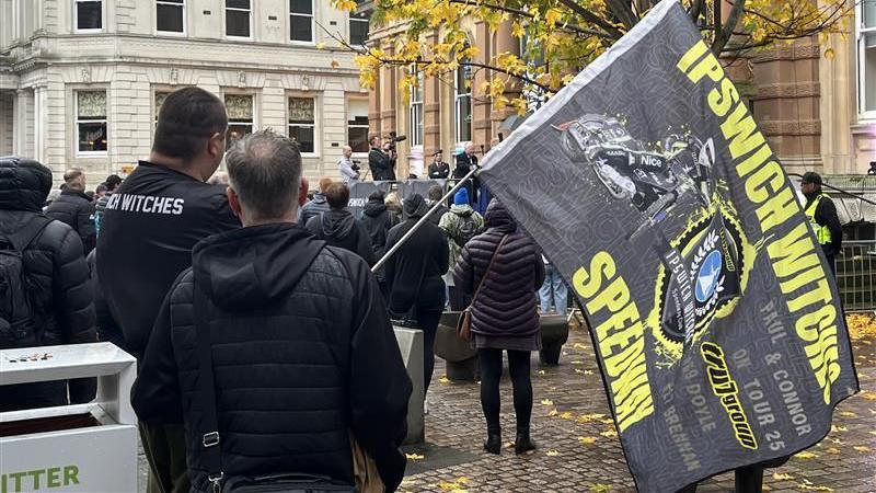A man in a black jacket with black hair holds a large black flag with the words in yellow letters, Ipswich Witches Speedway.