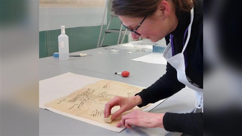 A woman with dark long hair and glasses wearing a dark jumper and a white apron. She is pushing a sponge onto a an old piece of paper with a sketch on it.